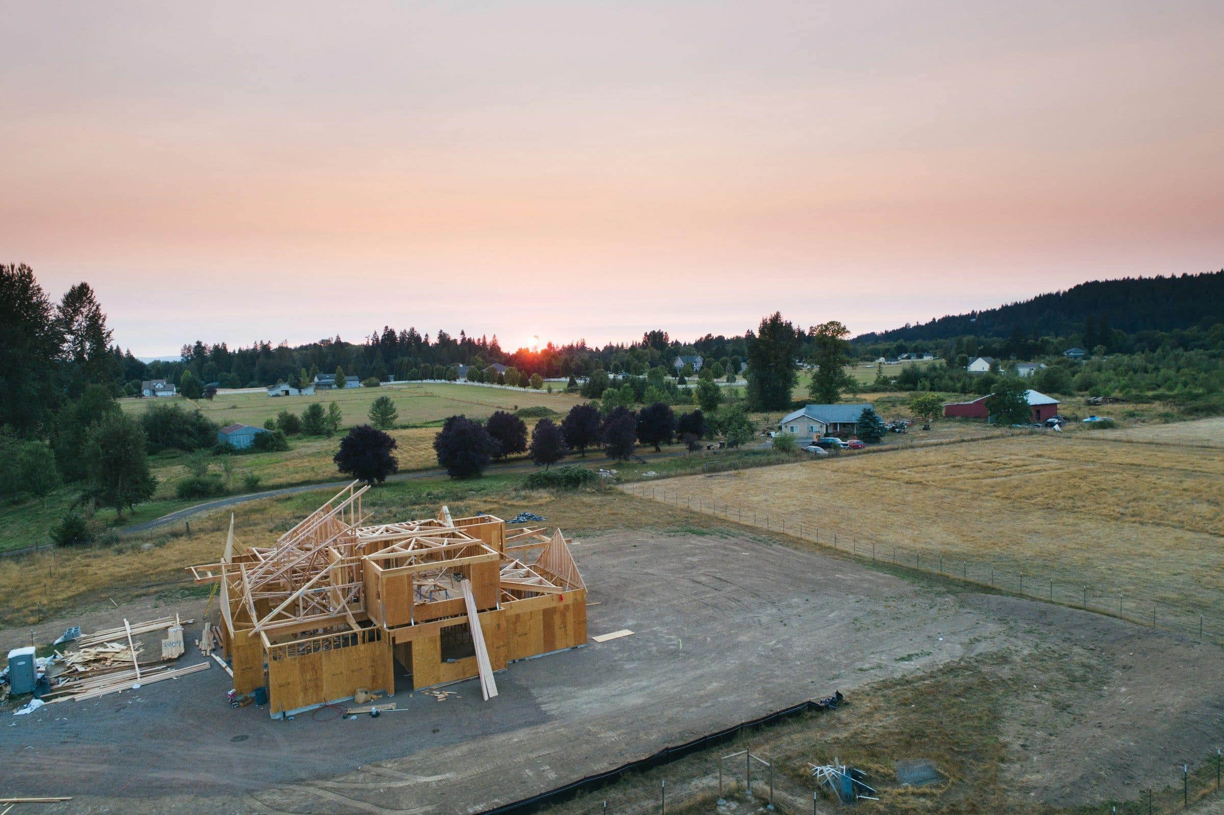 A timber-framed home under construction at sunset, set in open countryside